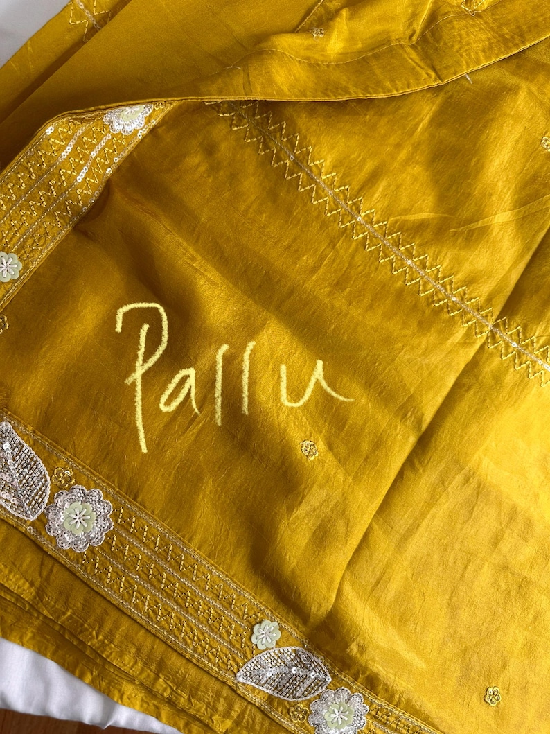 May include: Mustard yellow fabric with the word "Pallu" in a handwritten style. The fabric has decorative borders with floral and leaf designs, and a stitched pattern. The fabric is folded and appears to be silk or a similar material.