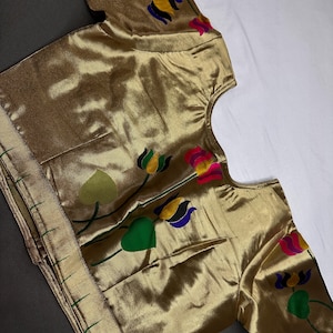 May include: A gold-colored blouse with short sleeves. The blouse features colorful floral embroidery in green, yellow, blue, and pink. The fabric has a subtle sheen, and the edges are trimmed with a gold border.