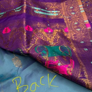 Op de afbeelding: Close-up van een levendig textiel met een rijke paarse tint, versierd met ingewikkelde gouden patronen en accenten van teal en roze. De stof heeft sierlijke ontwerpen en een contrasterende lichtblauwe basis met het woord "Back" in geel.