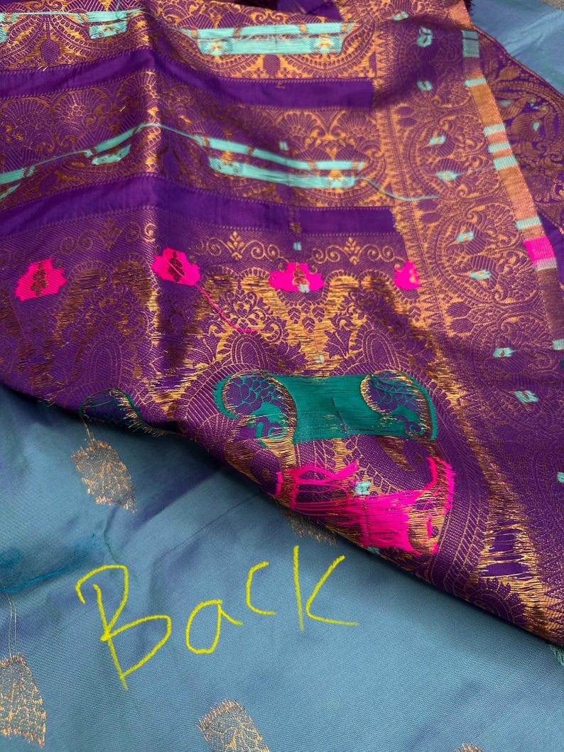 Op de afbeelding: Close-up van een levendig textiel met een rijke paarse tint, versierd met ingewikkelde gouden patronen en accenten van teal en roze. De stof heeft sierlijke ontwerpen en een contrasterende lichtblauwe basis met het woord "Back" in geel.