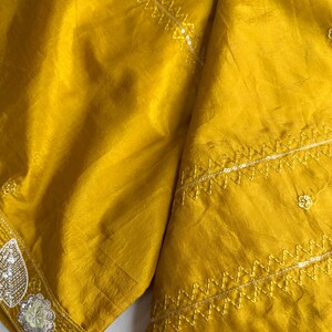 May include: Mustard yellow fabric with intricate embroidery and sequin detailing. The fabric has a subtle sheen and is folded, showcasing the texture and design. The edges are adorned with delicate embellishments, and a tassel hangs from the corner.