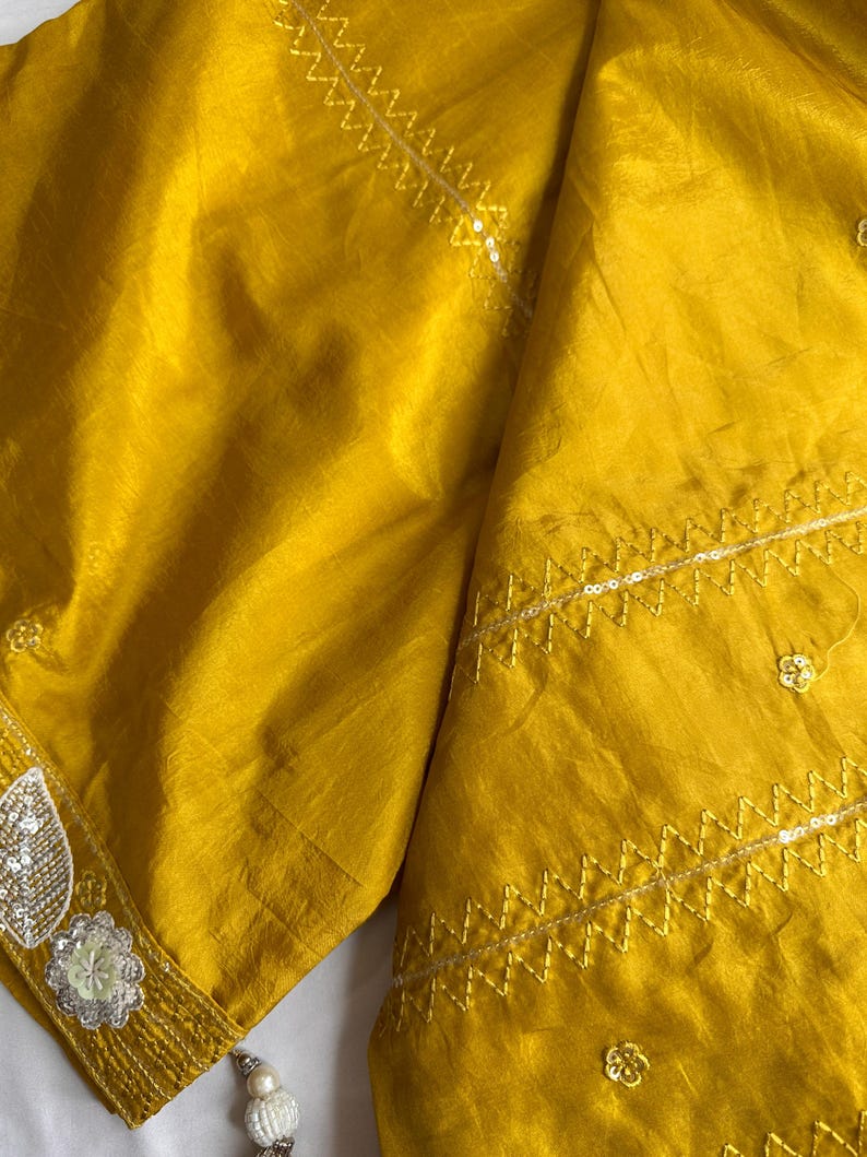 May include: Mustard yellow fabric with intricate embroidery and sequin detailing. The fabric has a subtle sheen and is folded, showcasing the texture and design. The edges are adorned with delicate embellishments, and a tassel hangs from the corner.