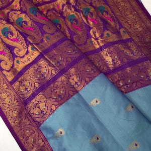 Op de afbeelding: Een zijden sari in paarse en blauwe tinten. Het heeft gouden paisley patronen met accenten van turquoise en roze. De rand is diep paars met gouden details. Het onderste gedeelte is effen lichtblauw. De stof is fijn geweven.