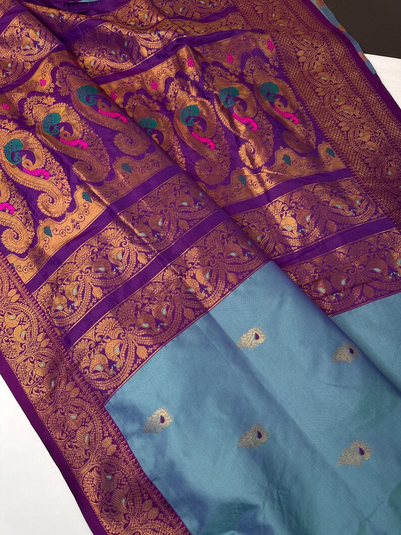 Op de afbeelding: Een zijden sari in paarse en blauwe tinten. Het heeft gouden paisley patronen met accenten van turquoise en roze. De rand is diep paars met gouden details. Het onderste gedeelte is effen lichtblauw. De stof is fijn geweven.