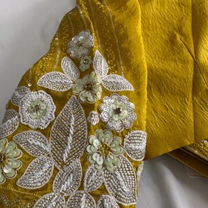 May include: Mustard yellow garment with intricate floral embroidery on the sleeve. The embroidery features white and light green sequins and beads, creating a textured, decorative design. The fabric has a subtle sheen.