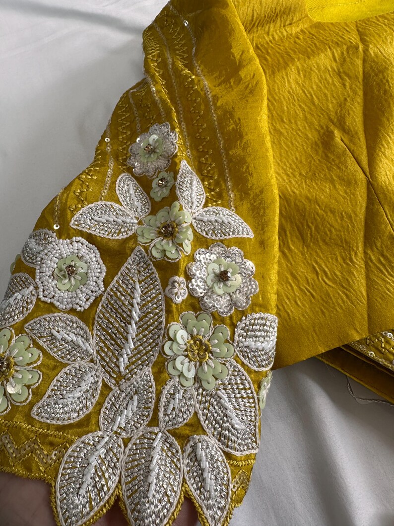 May include: Mustard yellow garment with intricate floral embroidery on the sleeve. The embroidery features white and light green sequins and beads, creating a textured, decorative design. The fabric has a subtle sheen.
