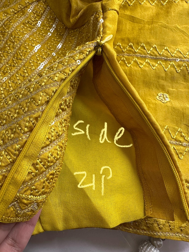 May include: Close-up of a mustard yellow garment with intricate embroidery and sequin detailing. The side zipper is visible, with the words "side zip" written in white. The fabric has a silky sheen.