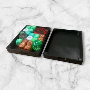 May include: A black dice box, open to reveal a collection of multi-coloured dice. The dice are numbered and include green, brown, white, and red dice. The box has a separate lid. The box is rectangular.