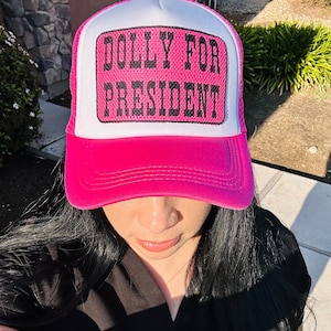 May include: A pink trucker hat with a white mesh back. The hat has a white patch with black text that reads "DOLLY FOR PRESIDENT".