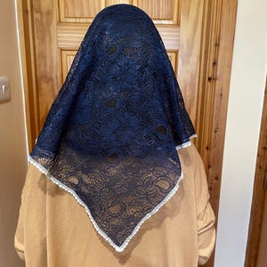 May include: A triangular head covering made of dark navy blue lace with a delicate floral pattern. The edges of the lace fabric are finished with a thin, white scalloped trim that outlines the entire perimeter of the triangular-shaped headpiece.