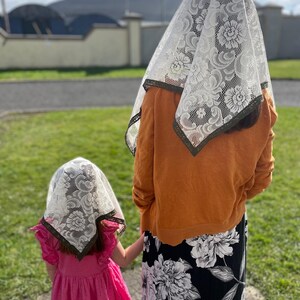 May include: Two people wearing white lace head coverings with floral patterns. One person is wearing an orange sweater and a black floral skirt. The other person is wearing a pink dress. They are standing on a sidewalk.