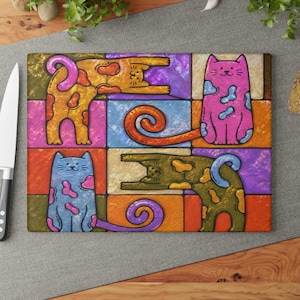 May include: A rectangular glass cutting board featuring a colorful grid pattern with four stylized cats. Two cats are sitting upright in pink and blue, while two cats are stretching horizontally in orange and olive green, all with spotted patterns.