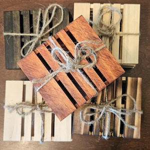 May include: A selection of wooden soap dishes in shades of brown, from dark to light. Each square dish has a slatted design and is tied with twine. The items are arranged on a wooden surface, creating a rustic aesthetic.