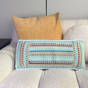 May include: A rectangular crochet pillow with a light blue, tan, and teal striped pattern. The pillow has a white scalloped edge and is placed on a white upholstered chair. A yellow pillow is behind it.