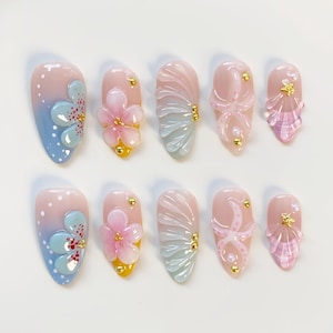 Könnte beinhalten: Ein Set aus zehn künstlichen Nägeln in verschiedenen Designs. Die Nägel zeigen einen Farbverlauf von Rosa und Blau, mit 3D-Blumen- und Muschelverzierungen. Einige Nägel haben goldene Akzente.