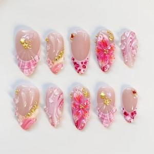 May include: A collection of ten almond-shaped artificial nails. Each nail has a nude base with pink and gold embellishments. Designs include floral patterns, glitter, and textured details. The nails are displayed on a white surface.