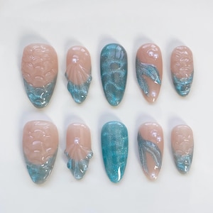 Könnte beinhalten: Ein Set von zehn künstlichen Nägeln mit Meerjungfrauen-Thema. Die Nägel zeigen eine Kombination aus hellrosa, türkis und silbernen Glitzerdesigns. Einige Nägel haben Muschel- und Perlenakzente, andere wellenartige Muster.