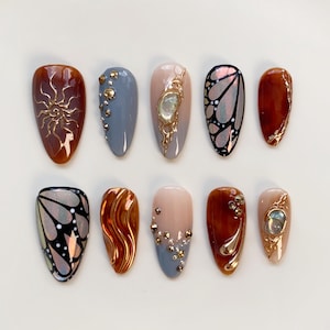 May include: Ten almond-shaped artificial nails with diverse designs. The nails showcase gold sunbursts, butterfly wing patterns, and embellishments with gold and silver rhinestones. The colour palette includes brown, grey, nude, and iridescent finishes.
