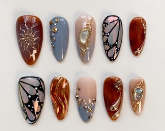 Puede incluir: Una colección de diez uñas artificiales en forma de almendra. Las uñas presentan varios diseños, incluyendo rayos de sol dorados, alas de mariposa y adornos con pedrería dorada y plateada. Los colores incluyen marrón, gris, nude y acabados iridiscentes.