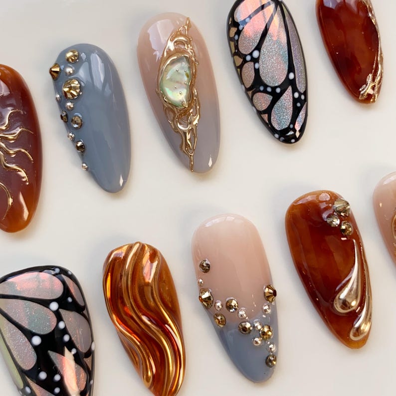 Puede incluir: Una colecci&oacute;n de u&ntilde;as artificiales en forma de almendra con varios dise&ntilde;os. Algunas u&ntilde;as presentan adornos dorados, patrones de mariposas iridiscentes y acentos de joyas. Los colores incluyen marr&oacute;n, gris, nude y un naranja brillante.