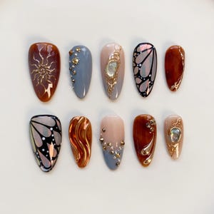 Puede incluir: Un conjunto de diez u&ntilde;as postizas en forma de almendra. Los dise&ntilde;os incluyen un sol, una mariposa y detalles dorados abstractos. Los colores var&iacute;an del marr&oacute;n, gris y nude al iridiscente y dorado, con adornos de gemas.