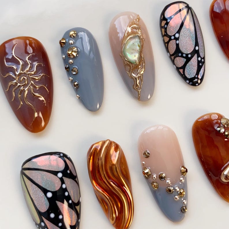 Puede incluir: Primer plano de una colecci&oacute;n de u&ntilde;as artificiales con diversos dise&ntilde;os. Las u&ntilde;as presentan colores marr&oacute;n, gris y nude, adornadas con adornos dorados, patrones de mariposas iridiscentes y dise&ntilde;os texturizados. Dispuestas sobre una superficie blanca.