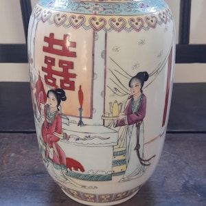 Puede incluir: Un jarrón de porcelana blanca con un diseño floral rosa, amarillo y verde. El jarrón presenta un carácter chino para "doble felicidad" y dos figuras con vestimenta tradicional.