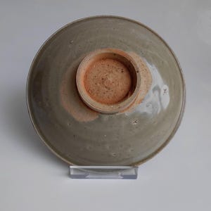 Peut inclure: Un bol en céramique vu par le dessous, présentant une glaçure brillante gris-vert clair avec de subtils motifs de craquelures. Le pied circulaire n'est pas émaillé, révélant une terre cuite brun-rouge texturée avec des imperfections.