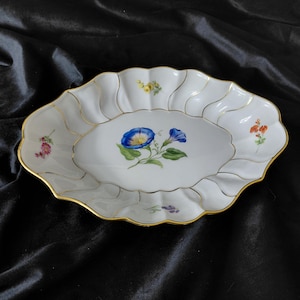 Op de afbeelding: Witte ovale serveerschaal met een geschulpte rand en gouden rand. De schaal heeft een centraal bloemmotief met blauwe bloemen en kleinere bloemaccenten in verschillende kleuren. De achtergrond is een donkere, getextureerde stof.