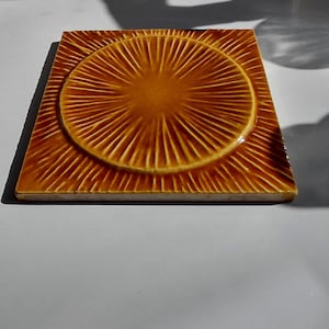 May include: A square ceramic tile with a glossy amber-brown glaze. The tile features a raised circular centre with radiating linear grooves that extend outward to the square edges, creating a sunburst pattern across the entire surface.