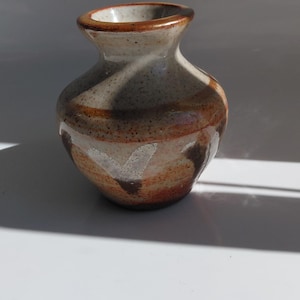 Puede incluir: Un pequeño jarrón de cerámica hecho a mano con cuerpo redondeado y cuello estrecho. El jarrón presenta una base beige moteada con bandas horizontales marrones y blancas. El borde es de color marrón más oscuro. El jarrón mide unos 10 cm de alto.