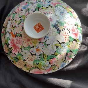 Puede incluir: Un cuenco redondo de cerámica blanca con un diseño floral en rosa, azul y verde. El cuenco tiene un pequeño centro elevado con un sello rojo. El cuenco está sobre una superficie negra.