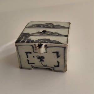 Puede incluir: Un pequeño recipiente cerámico rectangular con tapa, con un diseño de paisaje azul y blanco. El recipiente tiene un pico y elementos decorativos en la parte delantera. La estética general sugiere una influencia asiática tradicional.