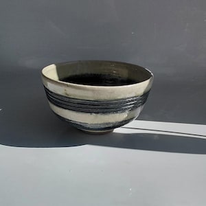Peut inclure: Un bol en céramique avec un extérieur crème et un intérieur sombre. Le bol présente trois bandes gris foncé autour du milieu. Le bol est posé sur un fond gris clair.