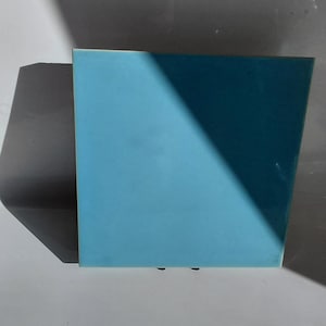 May include: A square, light blue object with a shadow cast across its surface. The object is set against a white and grey background. The object's colour is a solid, matte finish.