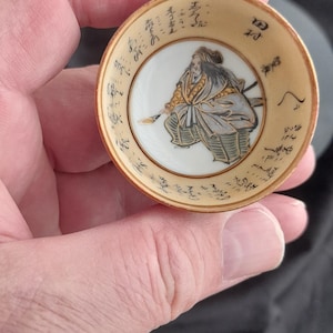 Può includere: Piccolo piatto rotondo in ceramica beige con un'illustrazione dettagliata di un guerriero samurai al centro. Il bordo presenta caratteri giapponesi. Il piatto è tenuto in mano.