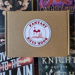 May include: A rectangular brown cardboard box featuring a circular white sticker on the front. The sticker displays the text Fantasy Book Club in dark pink lettering, surrounding a graphic of an open book, a castle, and a flying dragon.