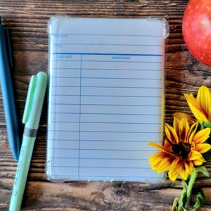 May include: A clear protective case with a library card design, blue pen, and a mint green pen rest on a wooden surface. A red apple and sunflowers with yellow and brown petals are also present. The library card design has lines for author, title, due date, and borrower.