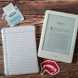 May include: A white Kindle e-reader displaying text from a book, with a clear case resembling lined paper. Decorative stickers include a typewriter and red vampire lips. The items are arranged on a wooden surface.
