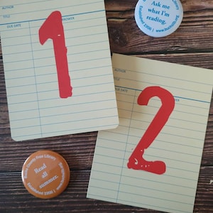 May include: Two library card-style notecards with the numbers 1 and 2 written in red. The notecards are on a wooden surface with two round buttons that say "Bucks County Free Library" and "Read all summer".