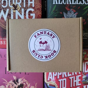 Puede incluir: Una caja de cartón marrón con una pegatina circular que dice "FANTASY BOOK CLUB". La pegatina presenta un diseño de dragón morado, castillo y libro abierto. La caja está rodeada de cubiertas de libros coloridas.