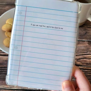 Pode incluir: Capa protetora transparente com design de papel de caderno. A capa apresenta linhas horizontais azuis, uma linha vertical vermelha e o texto "If you can read this, you're too close to me."