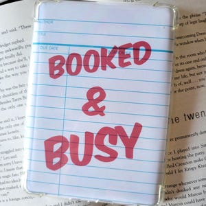 May include: A white phone case with a lined paper design and the words "BOOKED & BUSY" in red. The case has a clear border and is resting on open books.