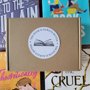 Puede incluir: Una caja de cartón marrón con una pegatina circular que dice "THE BOOK IS ALWAYS BETTER" alrededor de una ilustración de un libro abierto. La caja está rodeada de coloridas portadas de libros.