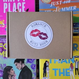 Può includere: Una scatola di cartone marrone con un adesivo bianco che dice "Romance Book Club" con un design di labbra rosse e viola. La scatola è circondata da copertine di libri con titoli come "Happy Place" e "Just for the Summer".