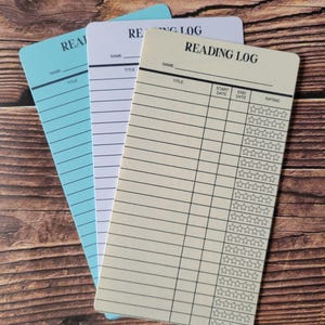 May include: Three reading log cards, one blue, one white, and one ivory. The ivory card has the words 'Reading Log' printed at the top. The cards have lines for writing the name, title, start date, end date, and rating of the book. The rating is indicated by five stars.
