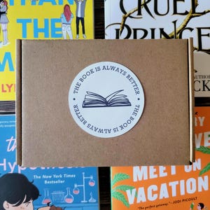 May include: A rectangular brown cardboard shipping box featuring a circular white sticker in the center. The sticker displays a black line drawing of an open book with the text "THE BOOK IS ALWAYS BETTER" printed in a circular pattern around it.