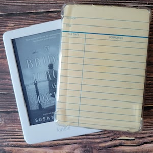 May include: A white Kindle e-reader displaying the book cover for A Bridge Across the Ocean by Susan Meissner, partially covered by a clear protective case featuring a vintage library checkout card design with lines for author, title, due date, and borrower.