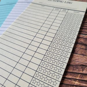 K&ouml;nnte beinhalten: Ein cremefarbenes Lesetagebuch mit einem schwarzen Raster und einer Spalte zur Bewertung von B&uuml;chern mit f&uuml;nf Sternen. Der Titel "Reading Log" ist oben auf der Seite gedruckt.