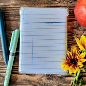 May include: A white library checkout card with blue lines and text reading AUTHOR, TITLE, DUE DATE, and BORROWER. The card is encased in a clear plastic protector. To the left are two pens, and to the right is a red apple and a yellow sunflower.